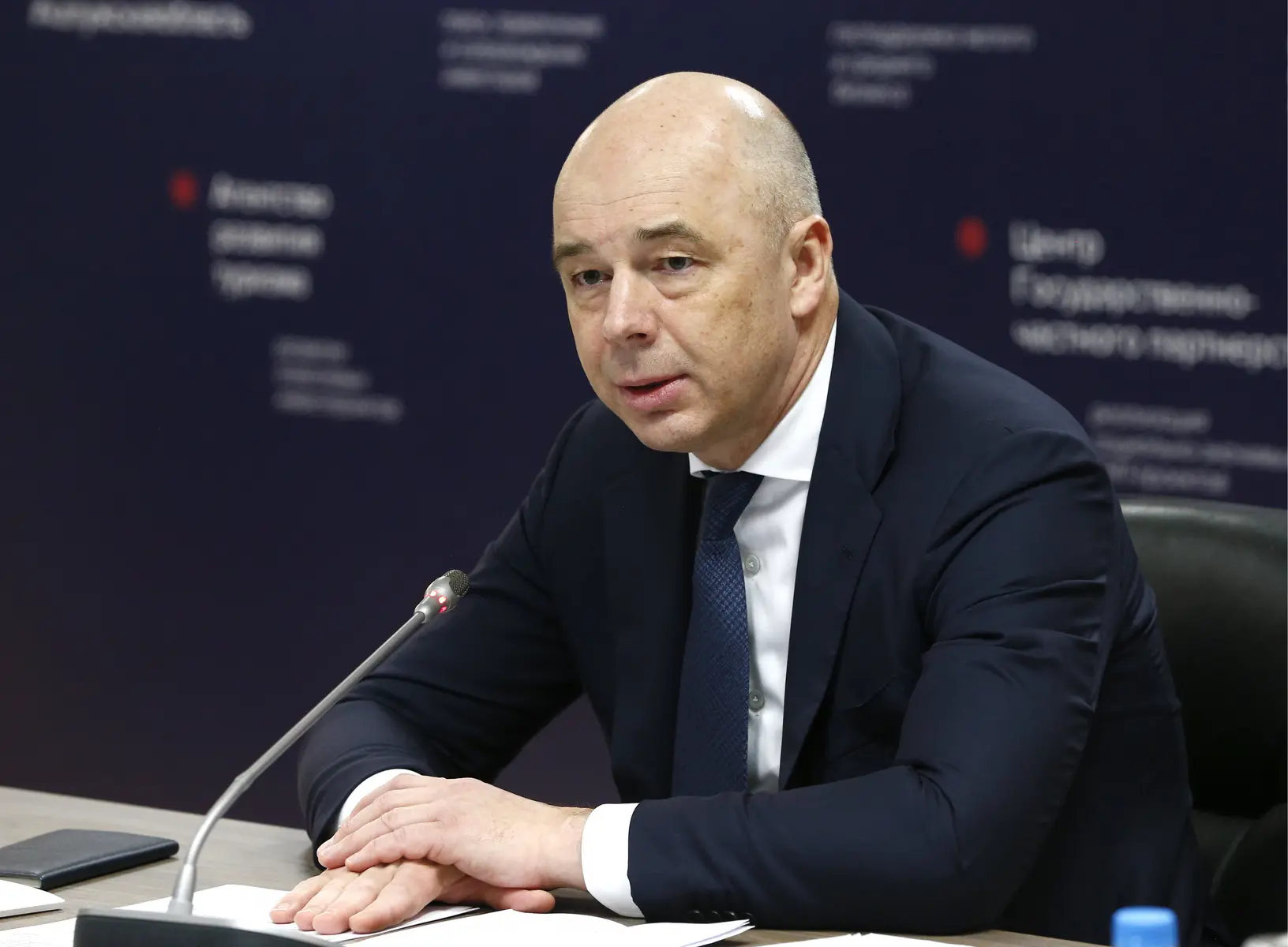 Anton Siluanov wearing a navy blue suit and tie, seated at a conference table with a microphone