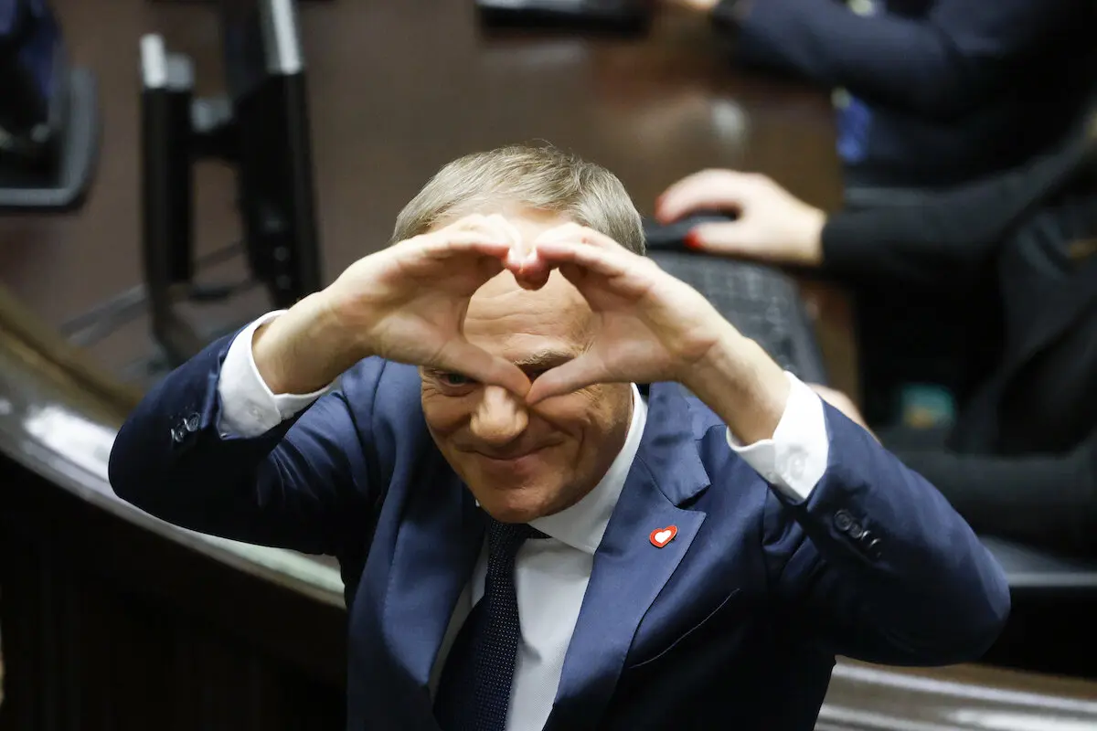 Donald Tusk in a blue suit and tie making a heart shape with his hands over his eyes.