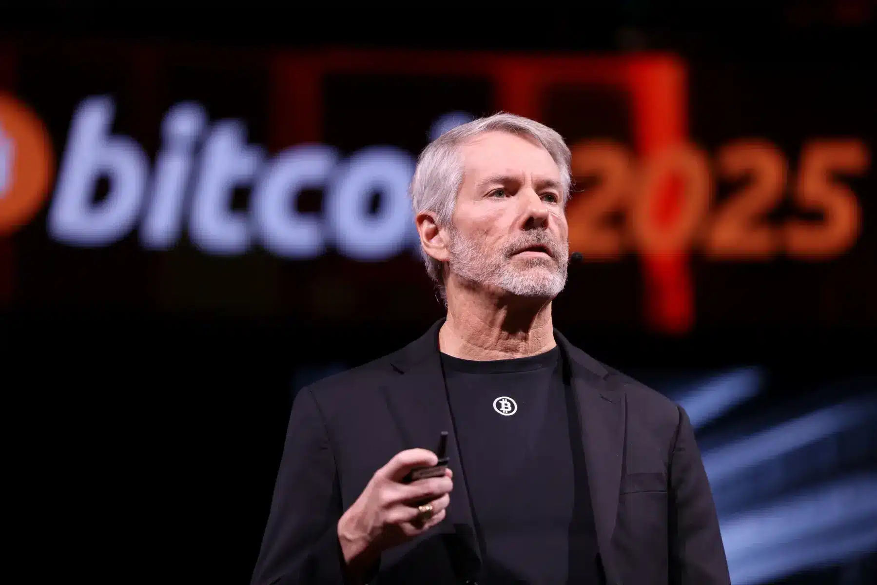 Michael Saylor wearing a Bitcoin logo t-shirt and blazer, speaking at a Bitcoin 2025 event