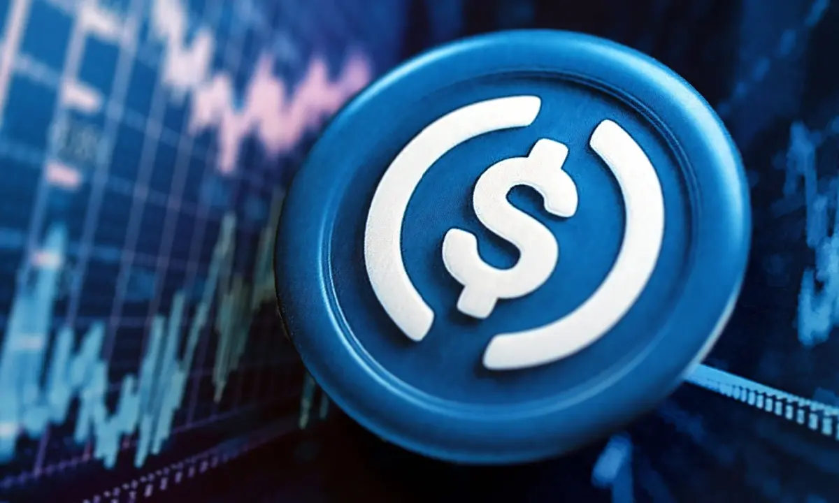 Close-up of a blue physical USDC token with a dollar sign and curved lines against a digital stock chart background