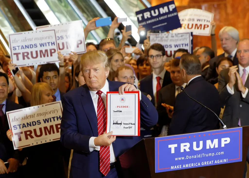 Donald Trump hält ein unterzeichnetes Dokument vor einem Podium und einer Menschenmenge mit Schildern.